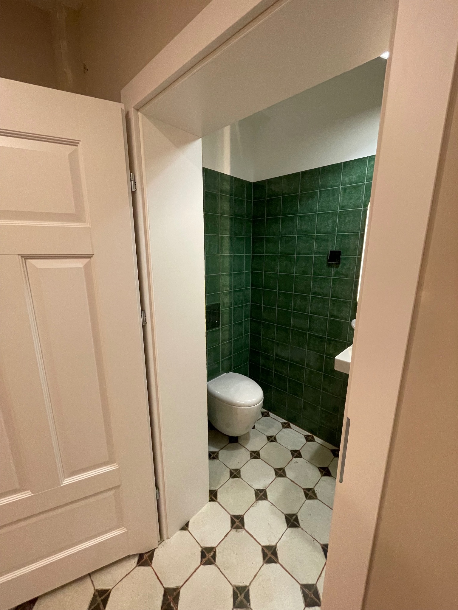 Otwarta łazienka z zielonymi płytkami ściennymi i wzorzystą podłogą. Widoczna toaleta i umywalka. Drzwi w kolorze kremowym.