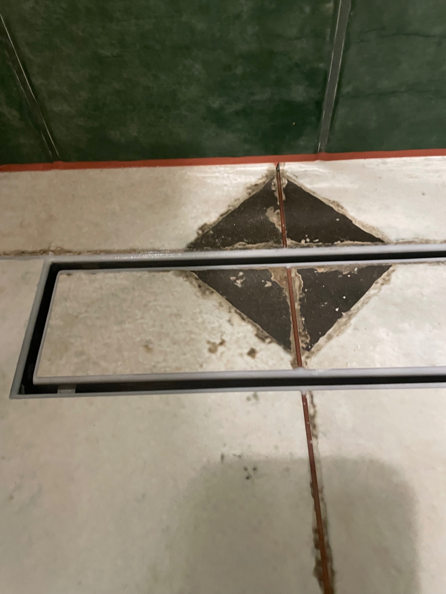 Ułożona podłoga w łazience z odpływem liniowym. Widoczne jasne płytki z ciemnymi fugami i zielona ściana w tle. Ujęcie z bliska.