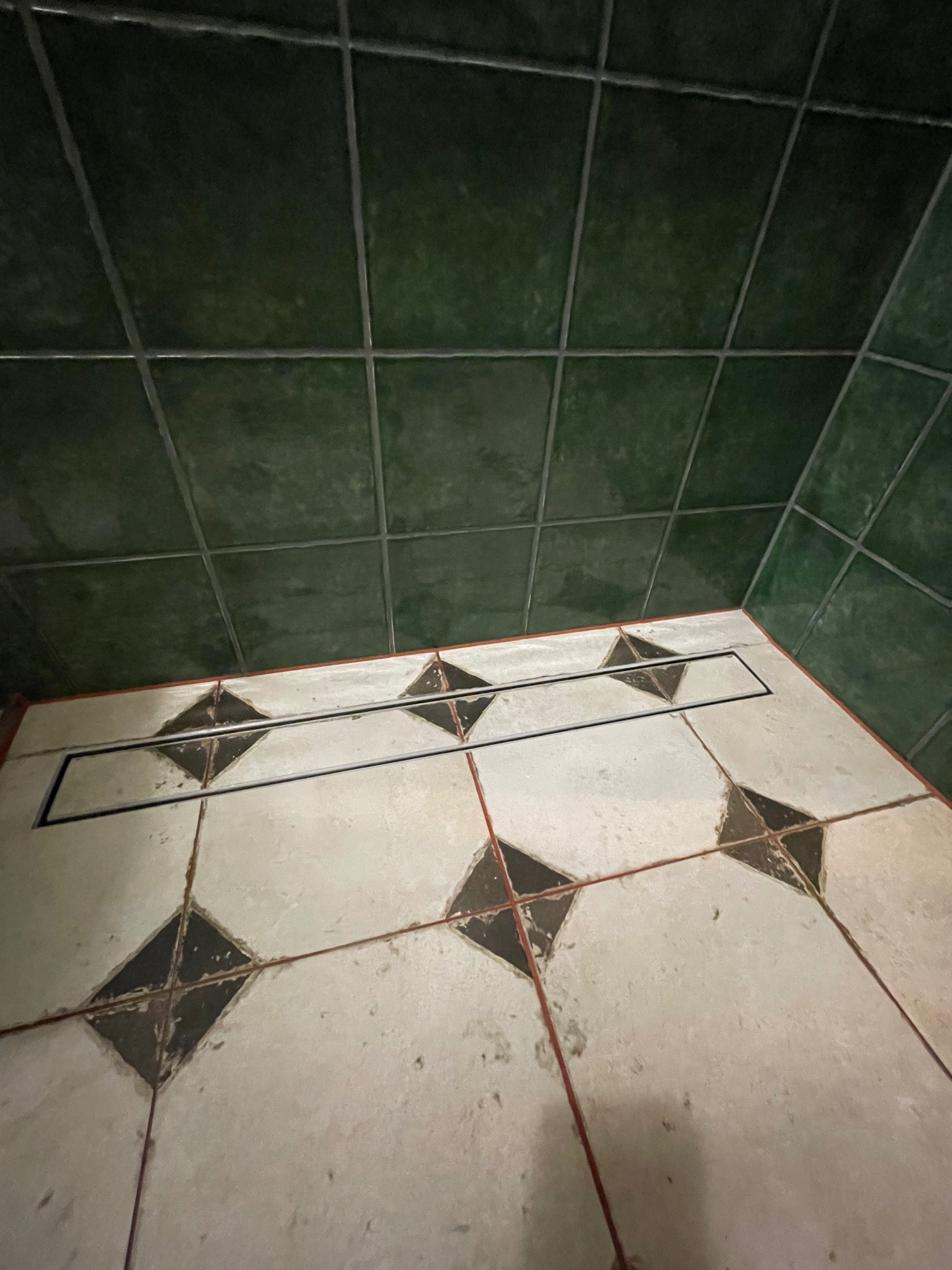 Ukończona łazienka z zielonymi płytkami na ścianach i białymi płytkami z czarnymi rombami na podłodze. Widoczny liniowy odpływ podłogowy.