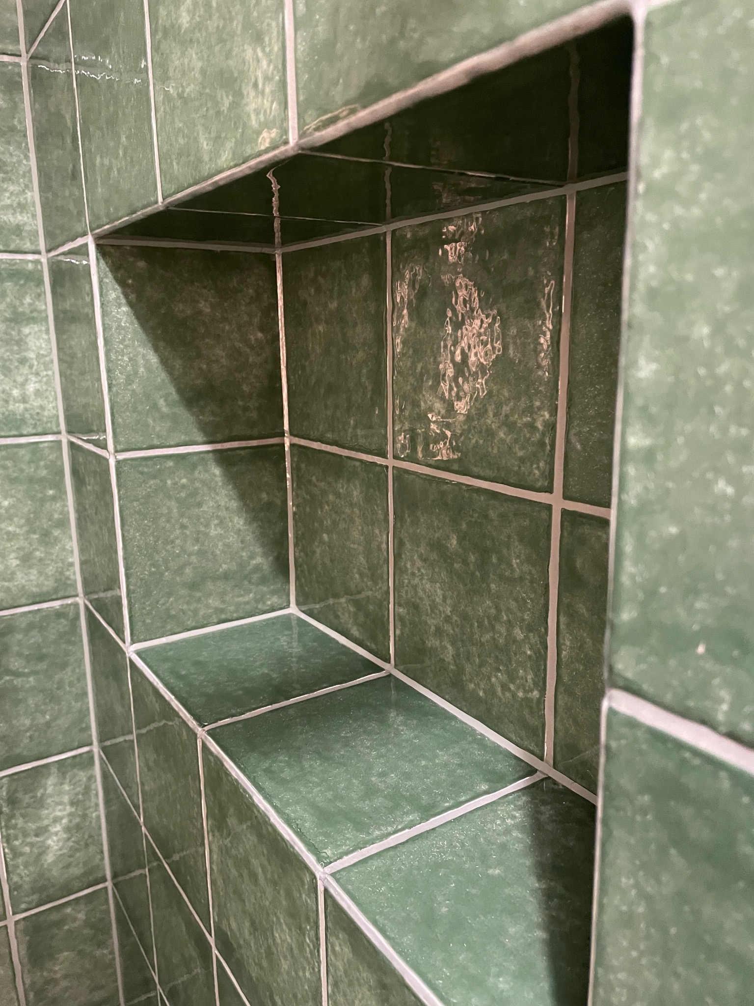 Narożna wnęka w ścianie wyłożonej zielonymi, kwadratowymi płytkami z jasną fugą. Widok z bliska, ukazujący detale wykończenia i fakturę płytek.