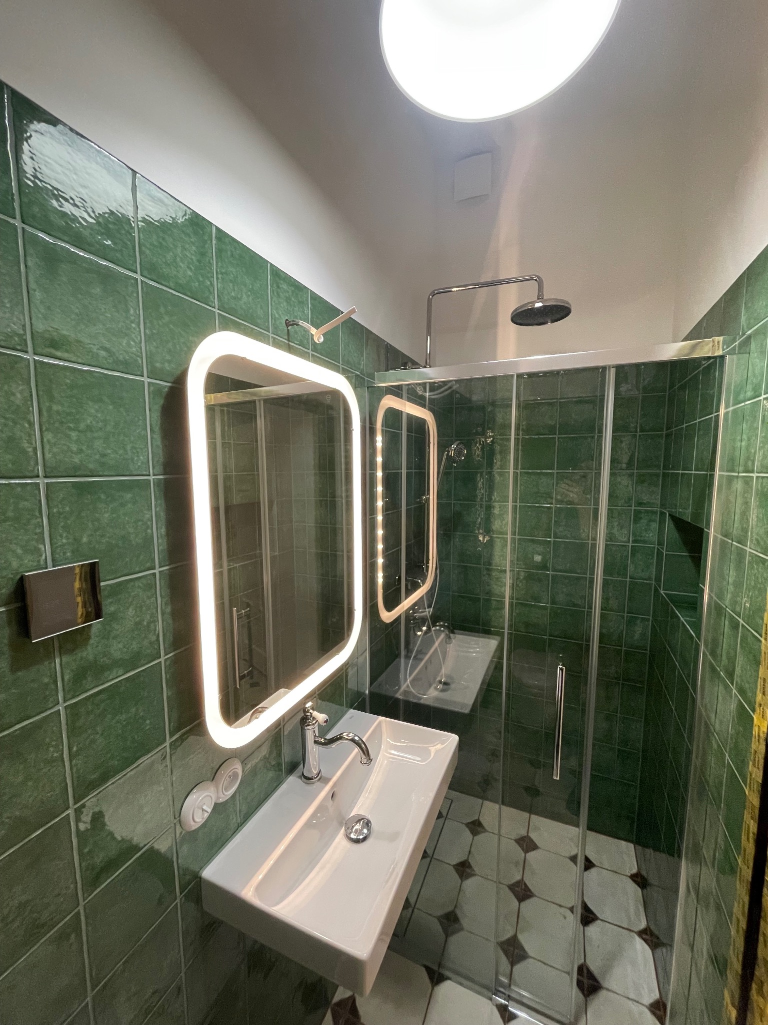 Nowoczesna łazienka z zielonymi, kwadratowymi płytkami, kabiną prysznicową ze szklanymi drzwiami i podświetlanym lustrem nad białą umywalką. Widoczne okrągłe okno w suficie.