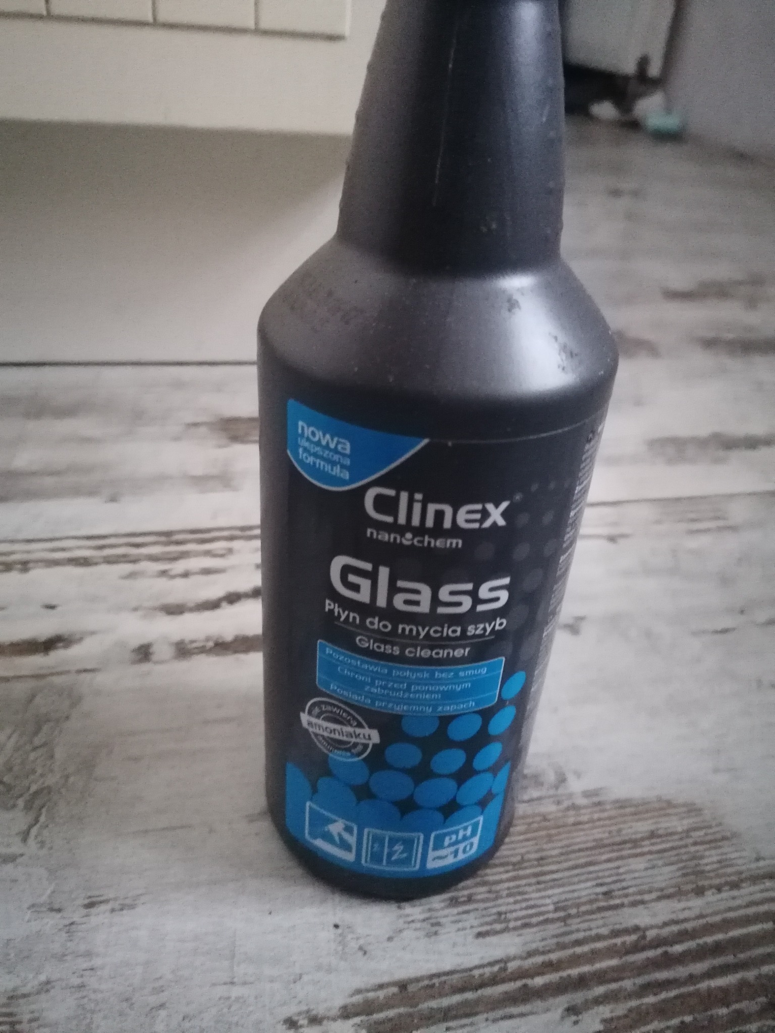 Butelka środka do mycia szyb Clinex Glass na drewnopodobnej powierzchni. Widoczne napisy 'nowa, ulepszona formuła', 'Glass Płyn do mycia szyb', oraz ikony informujące o właściwościach produktu.