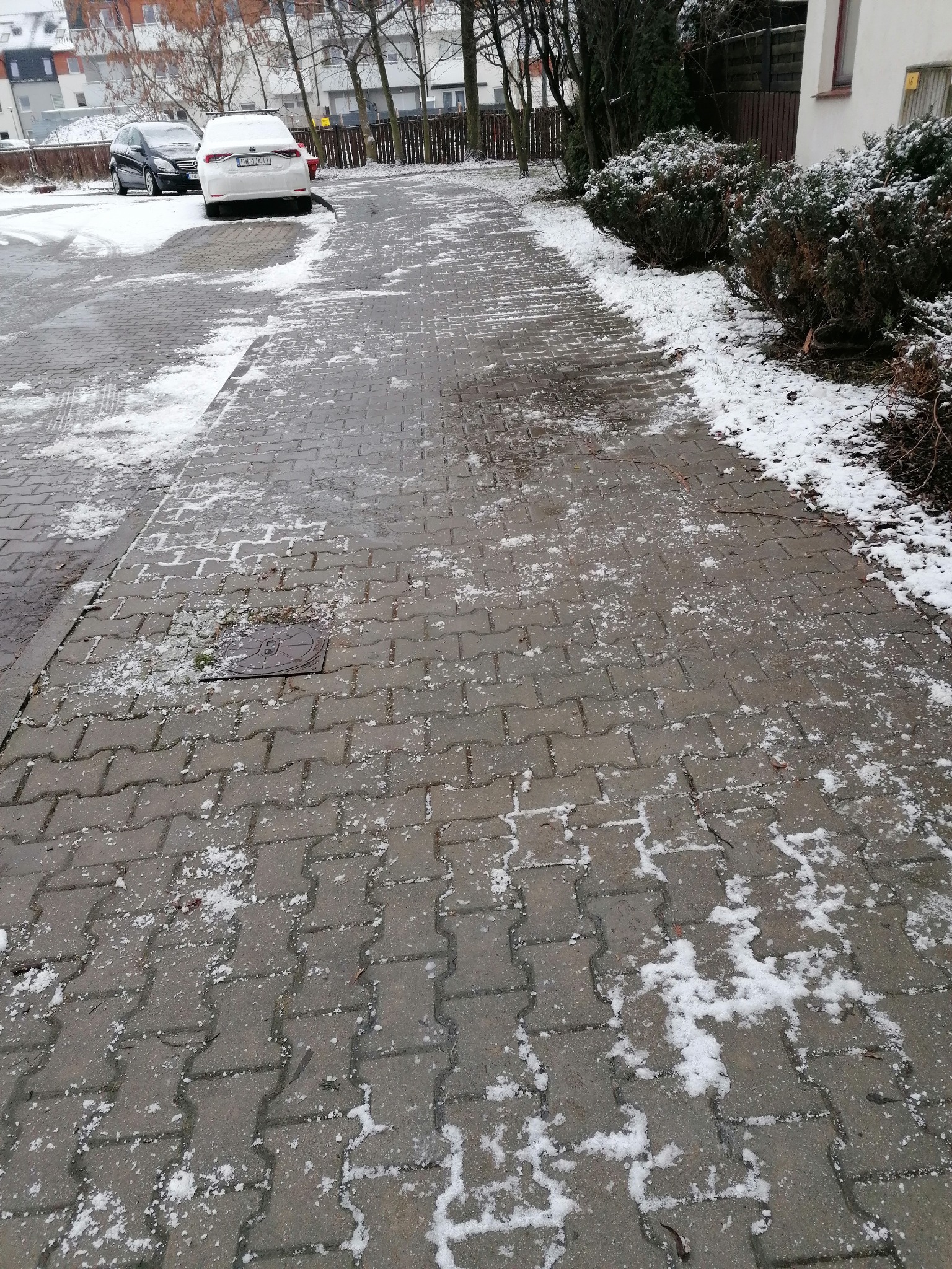 Kostka brukowa pokryta resztkami śniegu i lodu, widoczne ślady opon samochodowych, w tle zaparkowane auta i zabudowa mieszkalna.