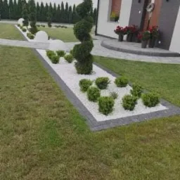 Nowoczesny ogród przydomowy z krętym drzewem, białym żwirem i kulistymi krzewami, z obrzeżami z szarej kostki brukowej, prowadzący do wejścia do domu z ciemnoszarą elewacją i dekoracyjnym wieńcem...