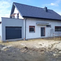 Nowoczesny dom z szarą elewacją i garażem w trakcie wykończenia, widoczne rusztowania i teren przygotowany pod zagospodarowanie.
