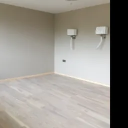 Jasne, drewniane panele podłogowe w pokoju z beżowymi ścianami, ozdobnym żyrandolem i dwoma kinkietami w foliowych osłonach.