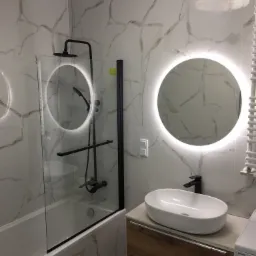 Nowoczesna łazienka z kabiną prysznicową typu walk-in z czarną armaturą, okrągłym lustrem z podświetleniem LED i umywalką nablatową na drewnianej szafce.