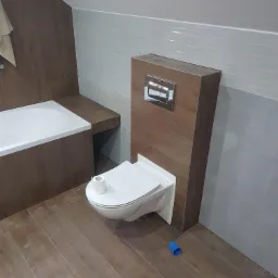 Nowoczesna łazienka w trakcie wykańczania z podwieszaną toaletą, drewnopodobnymi płytkami na podłodze i obudowie stelaża, oraz częściowo wykończonymi ścianami w odcieniach szarości i bieli.