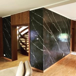 solidbud-remonty.pl - Salon z drewnianą podłogą i ścianami wykończonymi czarnym tynkiem imitującym marmur z białymi żyłkami, widoczne spiralne schody w tle.