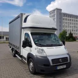 Biały Fiat Ducato z plandeką i windą załadowczą, zaparkowany na brukowanym placu, z budynkiem biurowym w tle i błękitnym niebem z chmurami.
