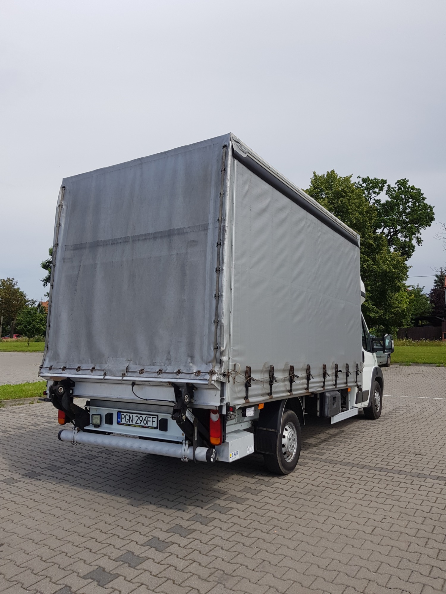 Tył białego samochodu dostawczego z plandeką na parkingu z kostki brukowej, widoczny numer rejestracyjny PGN 296FF.