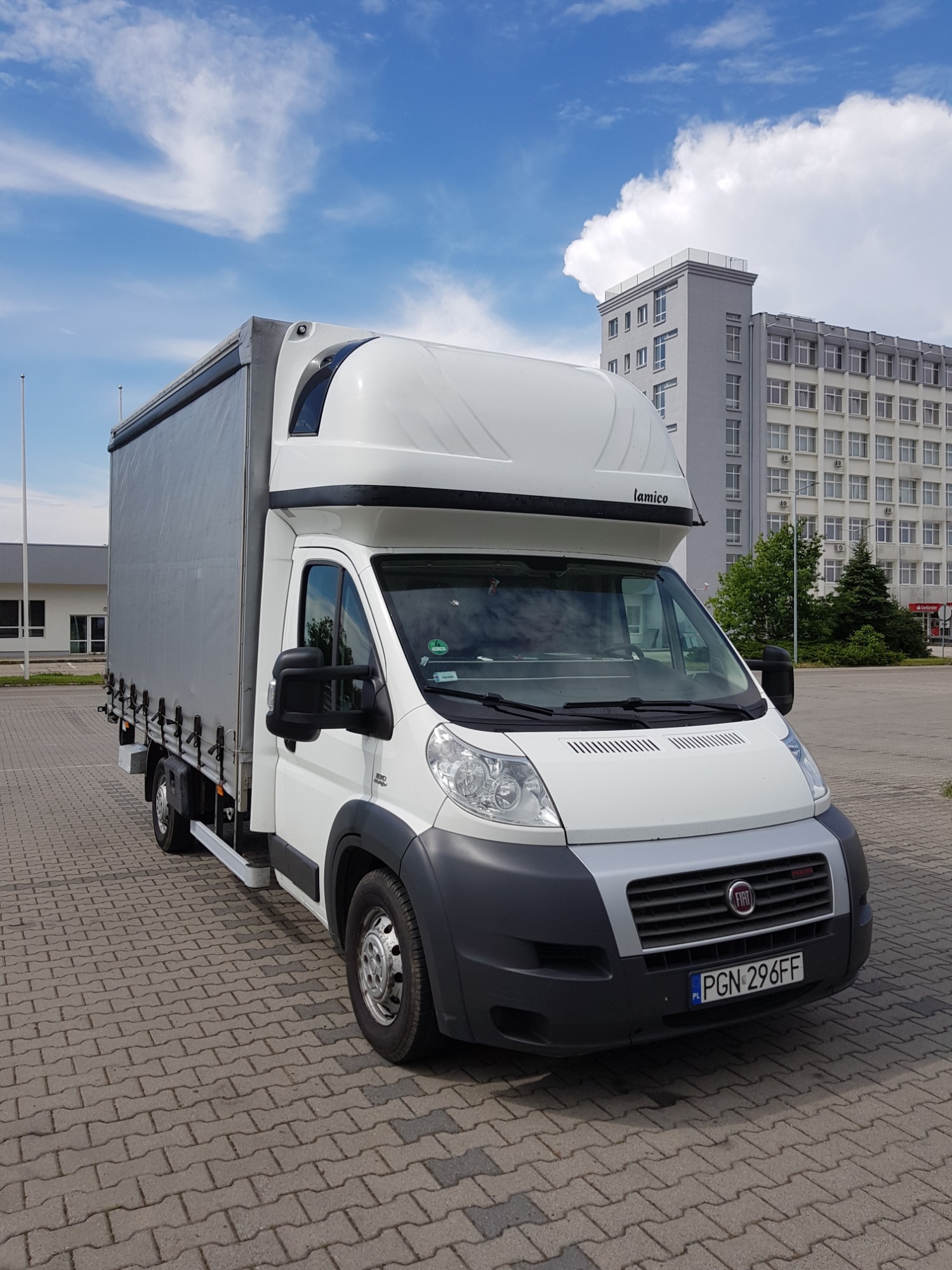Biały Fiat Ducato z plandeką i windą załadowczą, zaparkowany na brukowanym placu, z budynkiem biurowym w tle i błękitnym niebem z chmurami.