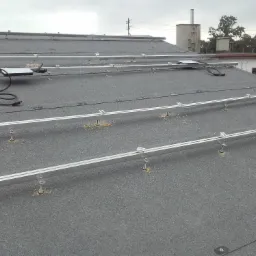 Montaż szyn pod panele słoneczne na bitumicznym dachu, widoczne elementy mocujące i kable, w tle budynek i drzewa przy pochmurnym niebie.