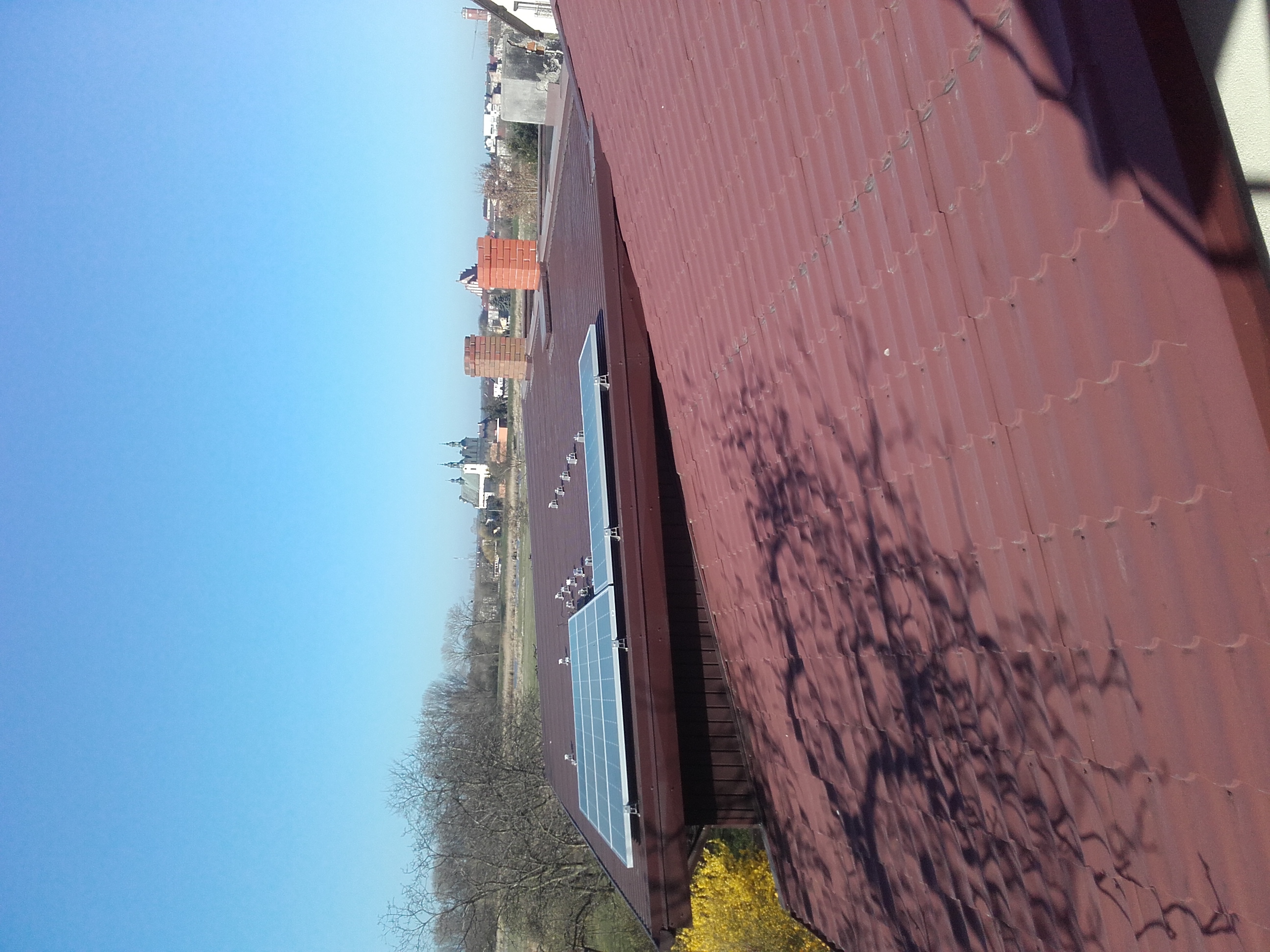 Panele słoneczne zamontowane na dachu z blachy trapezowej w odcieniach czerwieni, widok na okoliczną zabudowę z kominami i kościołem w tle, błękitne niebo.