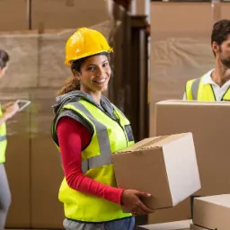 Pracownica w żółtym kasku i kamizelce odblaskowej uśmiecha się, niosąc karton w magazynie, w tle pracownicy z tabletem i kartonami.