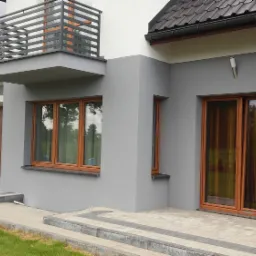 Nowoczesny, szary dom z drewnianymi oknami i balkonem z metalową balustradą, widok z zewnątrz na elewację i fragment podjazdu wyłożonego kostką.