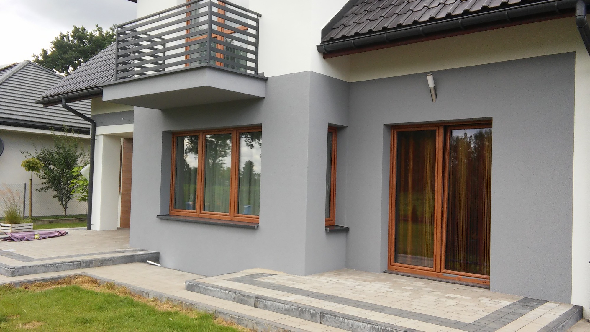Nowoczesny, szary dom z drewnianymi oknami i balkonem z metalową balustradą, widok z zewnątrz na elewację i fragment podjazdu wyłożonego kostką.