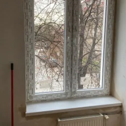 Nowe, białe, dwuskrzydłowe okno PCV marki Schüco z widokiem na drzewa bez liści, zamontowane w pokoju w trakcie remontu.