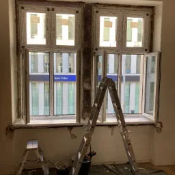 Montaż nowych okien PCV w starym budynku, widoczne okna z otwartymi skrzydłami, drabina, zabezpieczona podłoga i fragment elewacji z napisem Bank Polski