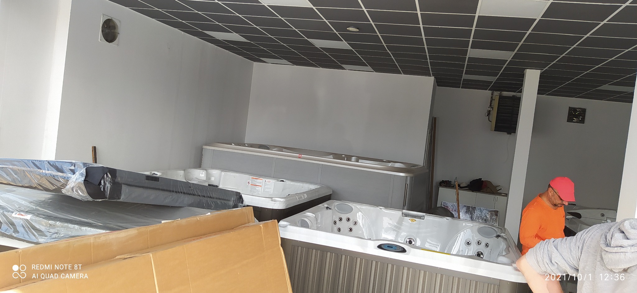 Wnętrze magazynu z kilkoma nowymi, zapakowanymi wannami z hydromasażem, pracownik w pomarańczowej koszulce i czerwonej czapce w tle.