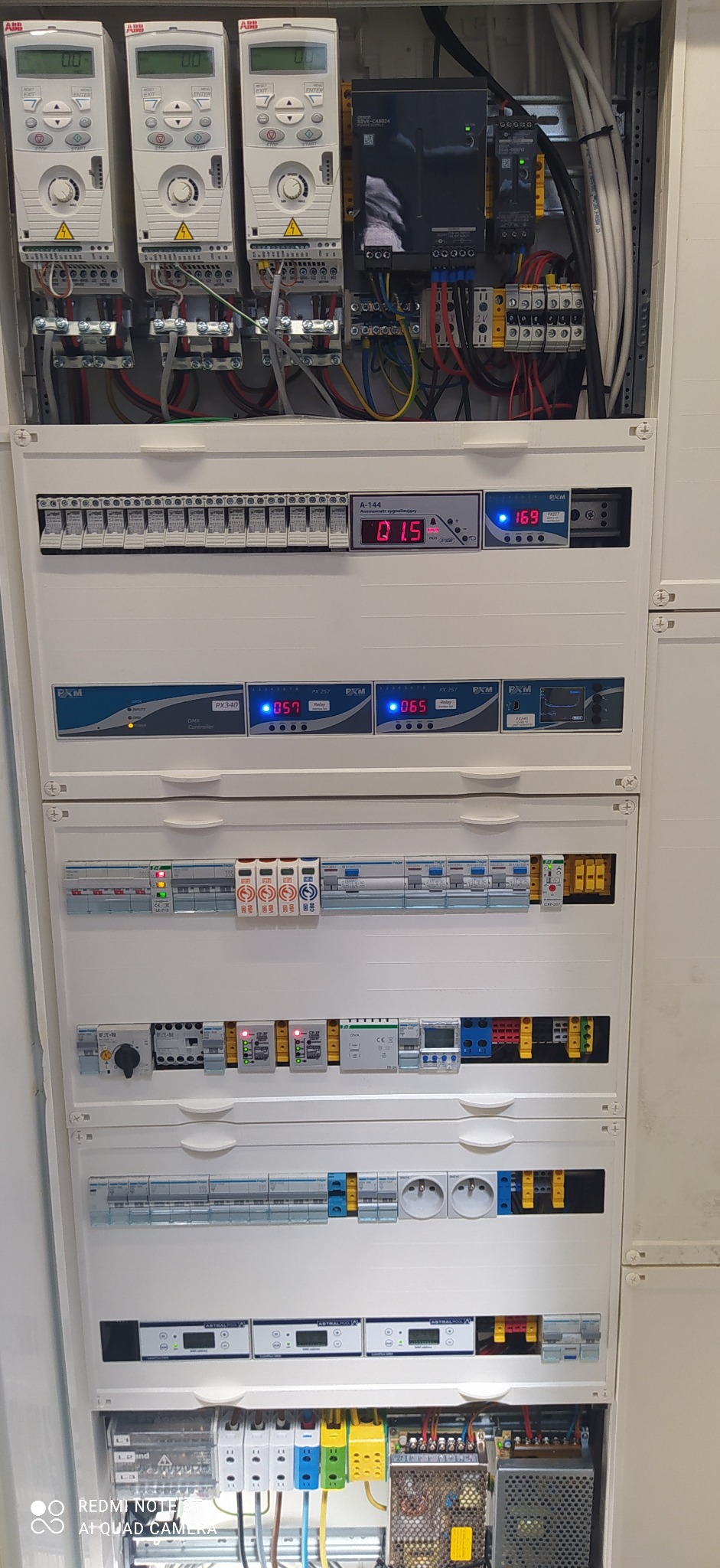 Rozdzielnia elektryczna z różnymi modułami sterującymi, bezpiecznikami i zasilaczami, widoczne kable i oznaczenia na urządzeniach.