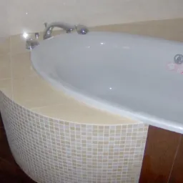 Zaokrąglona obudowa wanny wyłożona mozaiką w odcieniach beżu i bieli, z blatem z płytek ceramicznych w kolorze kremowym, widoczna armatura i fragment podłogi z paneli imitujących drewno.