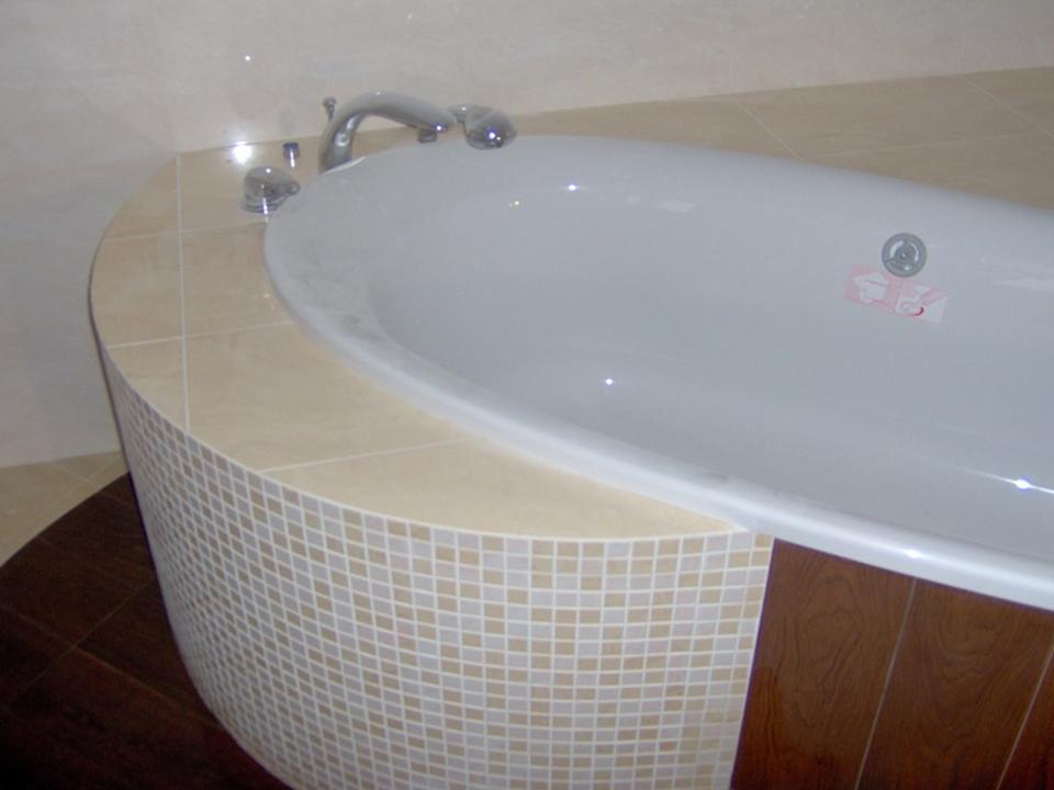 Zaokrąglona obudowa wanny wyłożona mozaiką w odcieniach beżu i bieli, z blatem z płytek ceramicznych w kolorze kremowym, widoczna armatura i fragment podłogi z paneli imitujących drewno.