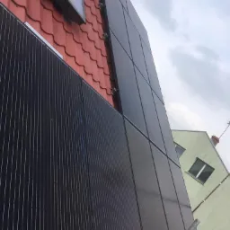 Montaż paneli słonecznych na dachu z czerwonej dachówki, widoczne uchwyty mocujące i fragment budynku w tle.