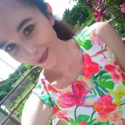 Portret uśmiechniętej kobiety w sukience w kwiaty, z widocznym naszyjnikiem i fragmentem balkonu w tle.