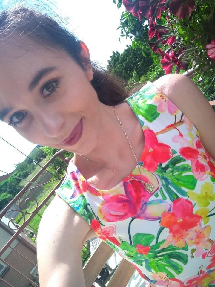 Portret uśmiechniętej kobiety w sukience w kwiaty, z widocznym naszyjnikiem i fragmentem balkonu w tle.