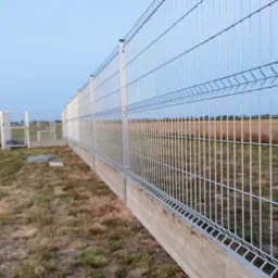 Długie, panelowe ogrodzenie z siatki z podmur&oacute;wką betonową, widoczne na tle pola i błękitnego nieba o zmierzchu, z fragmentem bramy w oddali.