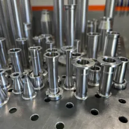 Precyzyjnie obrobione, srebrne elementy metalowe o różnych kształtach i rozmiarach, stojące na perforowanej płycie roboczej w warsztacie.