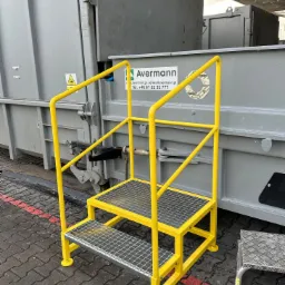 Żółte schody z poręczami, prawdopodobnie do kontenera, na zewnątrz, obok kontenera z logo Avermann.