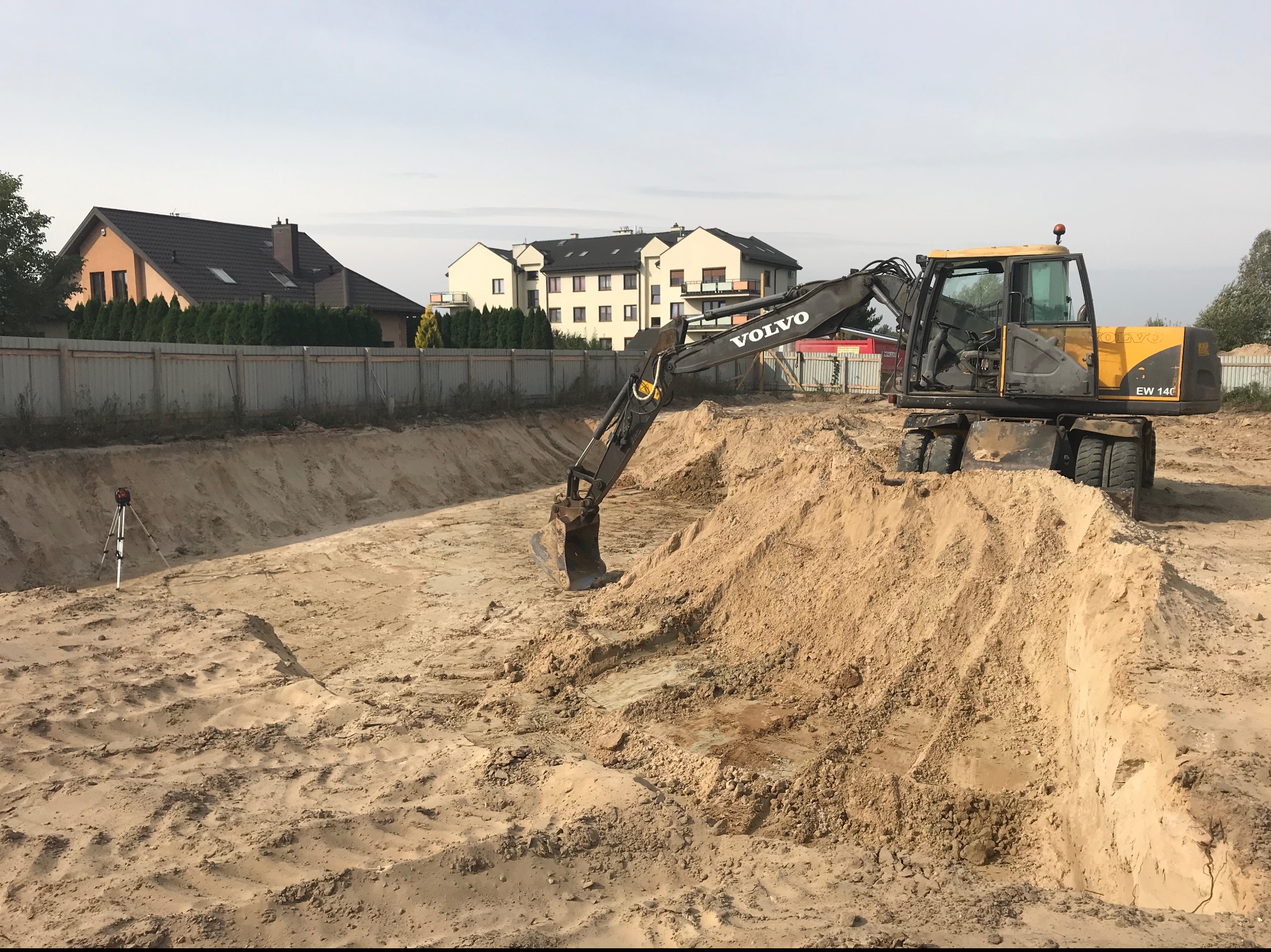 Żółta koparka kołowa Volvo EW 140 na placu budowy, z wykopem w piasku, w tle domy i ogrodzenie, widoczny niwelator laserowy na statywie.