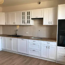 Jasna kuchnia z białymi frontami szafek, drewnianym blatem i czarnym zlewem, z zabudowanym piekarnikiem i mikrofalówką marki Amica.