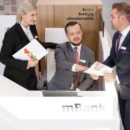 Wojciech Jastrzębski Fotografia Produktowa - Trzech pracowników banku w eleganckich strojach rozmawia przy ladzie, wręczając sobie dokumenty z logo banku i grafiką puzzli.