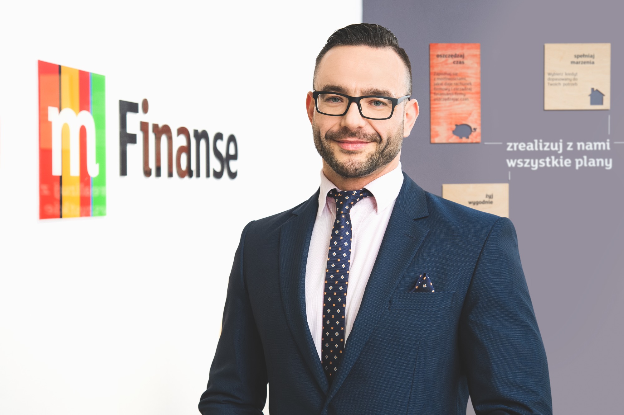 Portret uśmiechniętego mężczyzny w okularach i granatowym garniturze z poszetką, na tle biura firmy finansowej w Łodzi z logo i hasłami reklamowymi.