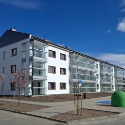 Nowy Dom Okna Sp. z o.o. - Nowoczesny, biały budynek wielorodzinny w Chodzieży z przeszklonymi balkonami i brązowymi ramami okiennymi, sfotografowany w słoneczny dzień na tle błękitnego nieba z nielicznymi obłokami,...