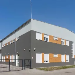 Nowoczesny budynek biurowy z szarą fasadą, białymi oknami i częściowo opuszczonymi roletami zewnętrznymi, widziany z niskiego kąta zza metalowego ogrodzenia.