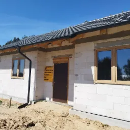 Surowy stan domu jednorodzinnego w trakcie budowy, z zamontowanymi oknami z drewnianymi ramami, widoczne rynny i tablica informacyjna budowy przy drzwiach.