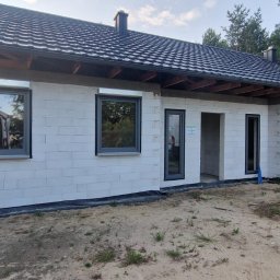 Nowy Dom Okna Sp. z o.o. - Dom w trakcie budowy z zamontowanymi szarymi oknami, białe ściany z bloczków, widoczne elementy dachu i podbitki, teren wokół w piasku, w tle las.