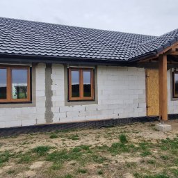Nowy Dom Okna Sp. z o.o. - Surowy stan domu jednorodzinnego w trakcie budowy, widoczne trzy okna z brązowymi ramami, ściany z białego betonu komórkowego, czarny dach z dachówki ceramicznej, oraz drewniana konstrukcja...