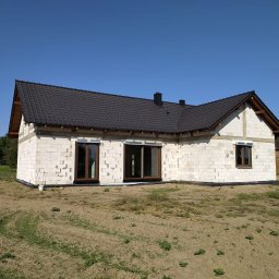 Nowy Dom Okna Sp. z o.o. - Surowy stan domu jednorodzinnego z białego bloczka, widoczne brązowe ramy okienne, czarny dach i przygotowany teren.