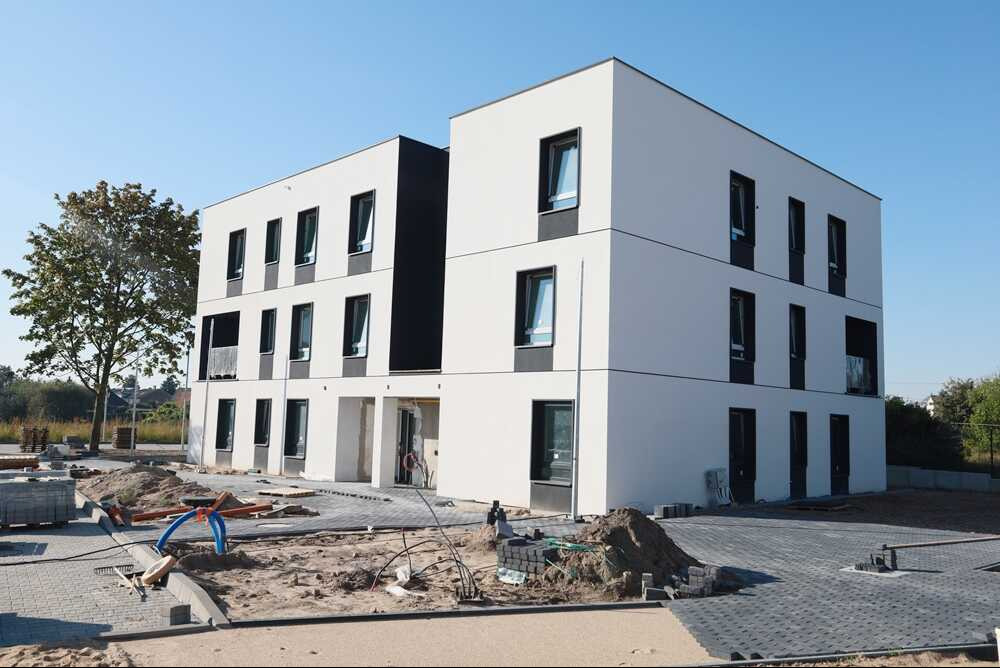 Nowoczesny, trzypiętrowy budynek mieszkalny w trakcie wykończenia, z widocznymi czarnymi ramami okiennymi i częściowo ułożoną kostką brukową wokół, na pierwszym planie widoczne materiały budowlane...
