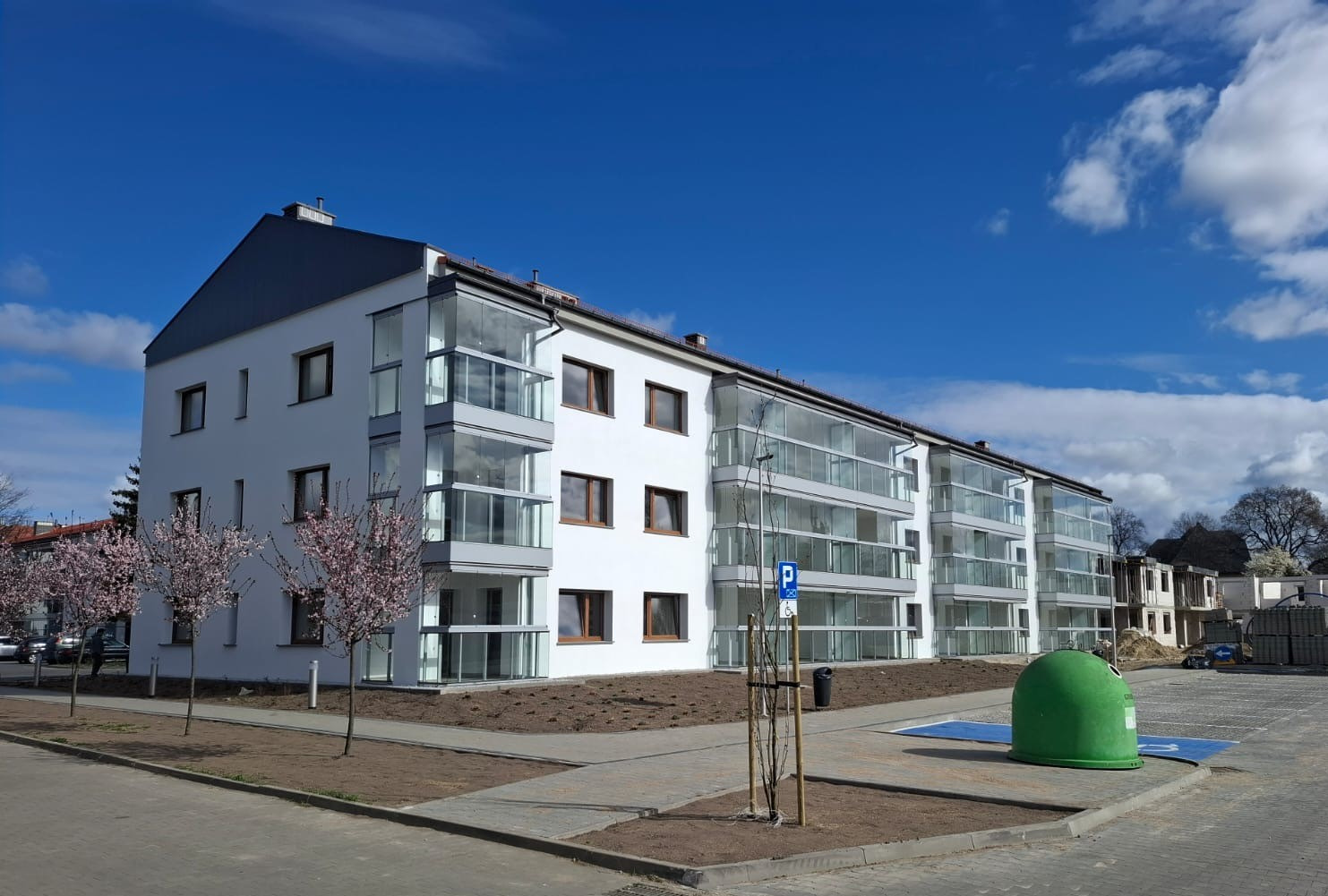 Nowoczesny, biały budynek wielorodzinny w Chodzieży z przeszklonymi balkonami i brązowymi ramami okiennymi, sfotografowany w słoneczny dzień na tle błękitnego nieba z nielicznymi obłokami,...