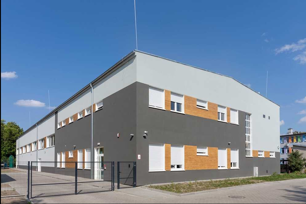 Nowoczesny budynek biurowy z szarą fasadą, białymi oknami i częściowo opuszczonymi roletami zewnętrznymi, widziany z niskiego kąta zza metalowego ogrodzenia.