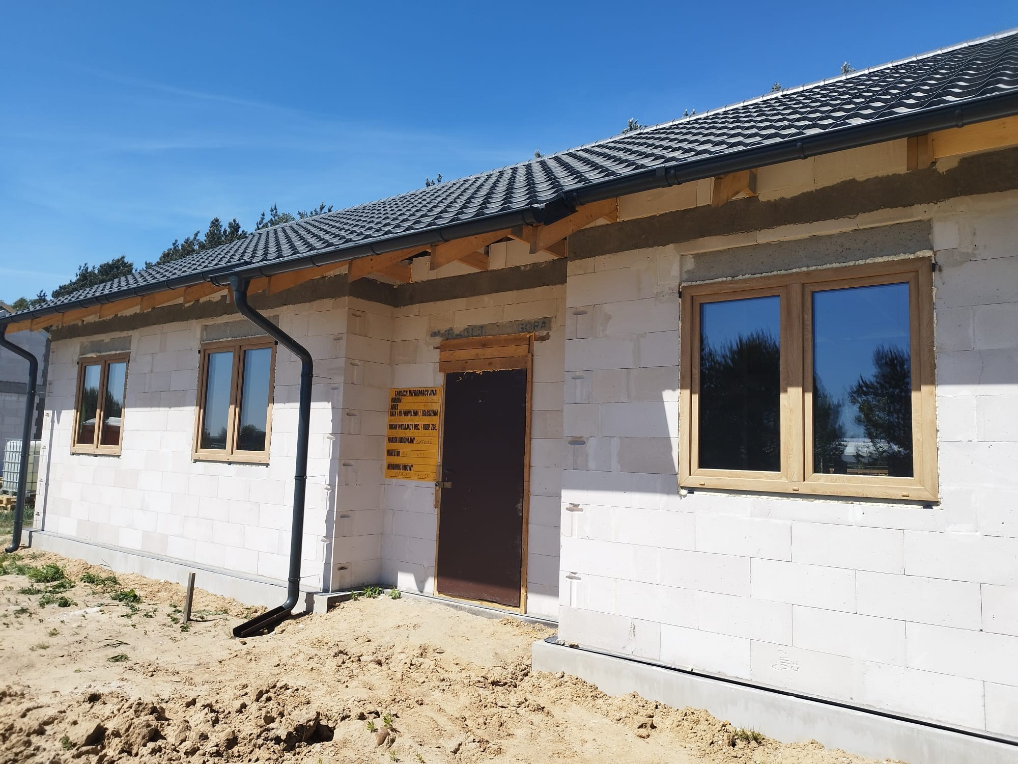 Surowy stan domu jednorodzinnego w trakcie budowy, z zamontowanymi oknami z drewnianymi ramami, widoczne rynny i tablica informacyjna budowy przy drzwiach.