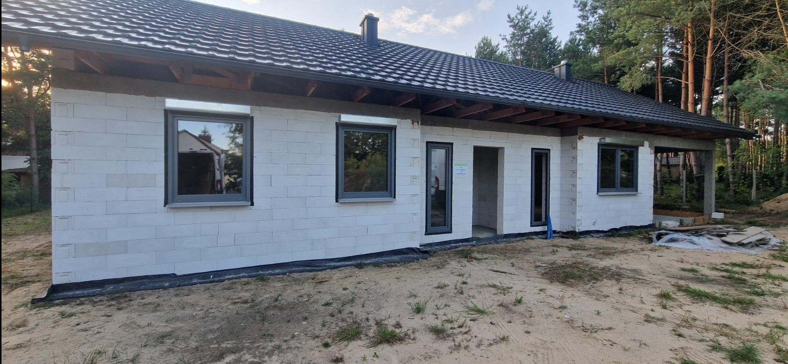 Dom w trakcie budowy z zamontowanymi szarymi oknami, białe ściany z bloczków, widoczne elementy dachu i podbitki, teren wokół w piasku, w tle las.
