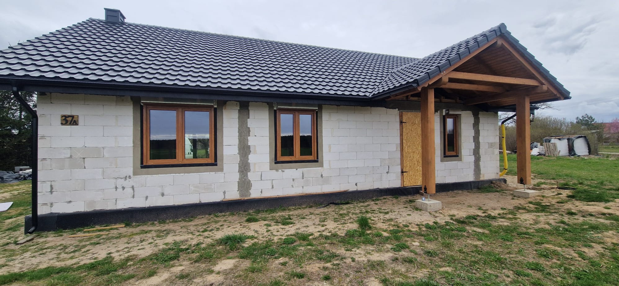 Surowy stan domu jednorodzinnego w trakcie budowy, widoczne trzy okna z brązowymi ramami, ściany z białego betonu komórkowego, czarny dach z dachówki ceramicznej, oraz drewniana konstrukcja...