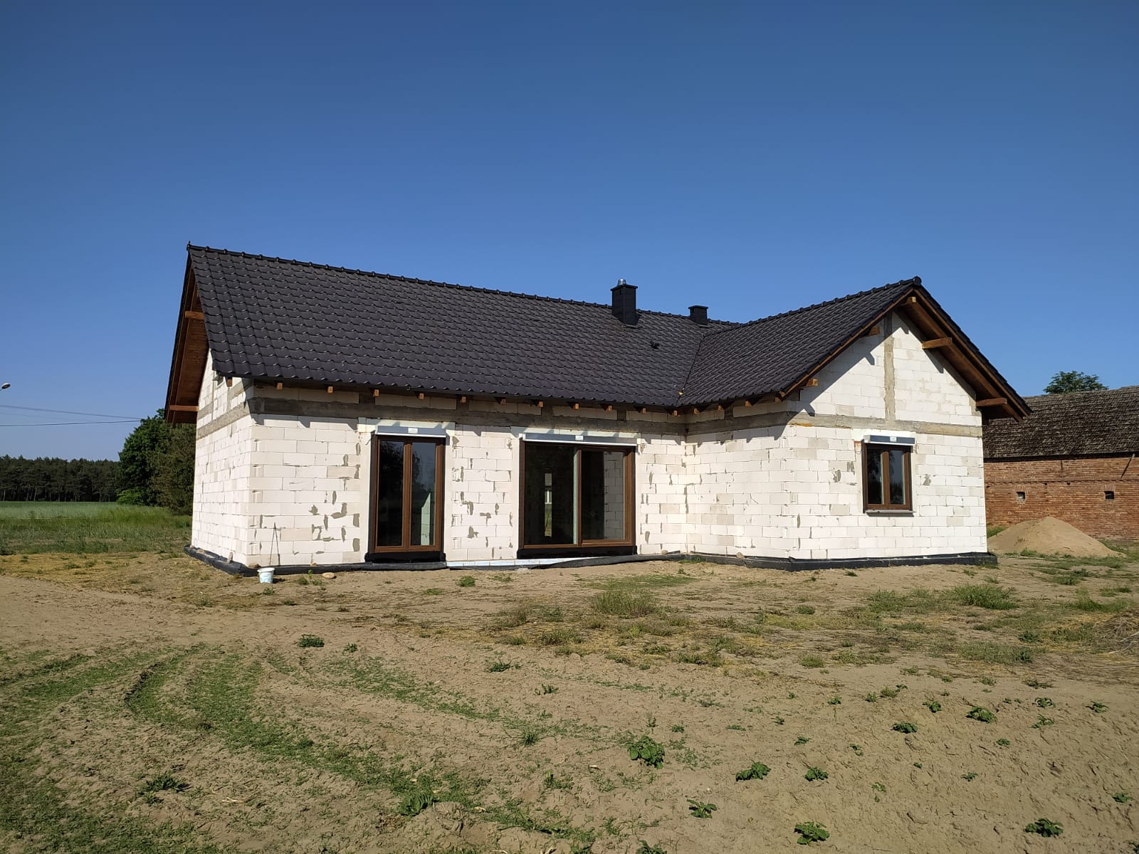 Surowy stan domu jednorodzinnego z białego bloczka, widoczne brązowe ramy okienne, czarny dach i przygotowany teren.